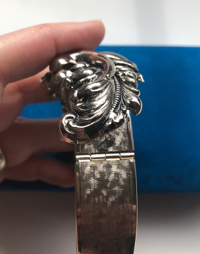 K&ouml;nnte beinhalten: Ein silbernes Armband mit einem dekorativen Blumenmuster. Das Armband ist mit einem Scharnier versehen und hat eine strukturierte Oberfl&auml;che.