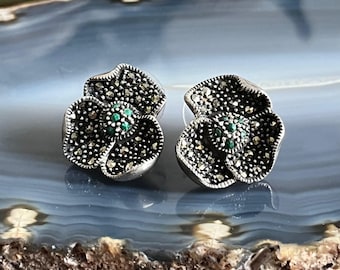 May include: A pair of silver flower-shaped earrings with green gemstones in the center. The earrings are made of marcasite.