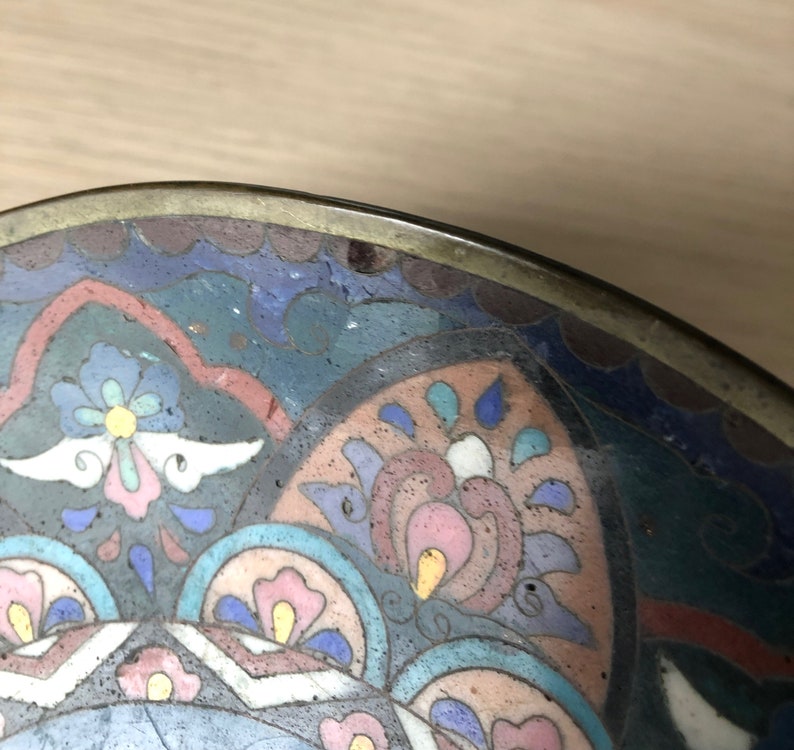 May include: A round, brass bowl with a colorful enamel design. The design features a floral pattern in shades of blue, pink, white, and yellow.