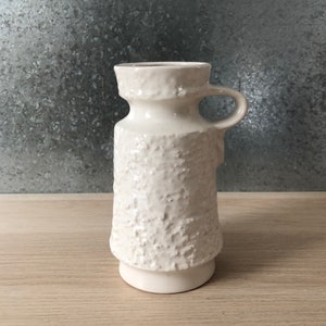 May include: White ceramic vase with a textured surface and a handle. The vase has a rounded base and a slightly flared top.