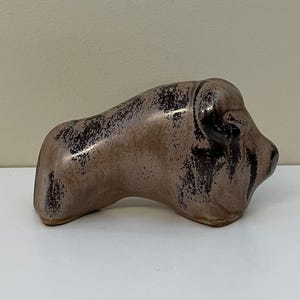 May include: A ceramic figurine of a bison in a seated position. The sculpture is primarily a muted brown color with black accents, giving it a rustic appearance. The bison's head is turned slightly to the right.