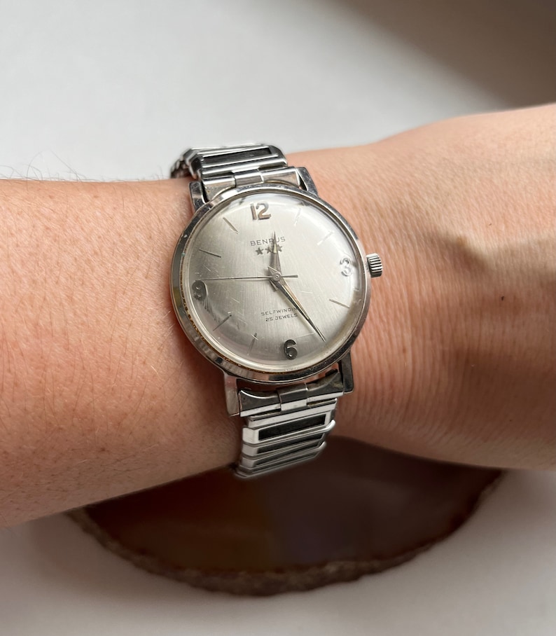May include: A silver wristwatch with a white face and black hands. The watch has a silver band and says "Benrus * * * Selfwinding 25 Jewels" on the face.
