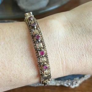 Könnte beinhalten: Ein goldenes Armreif-Armband mit einem dekorativen Design mit kleinen rosafarbenen Edelsteinen.