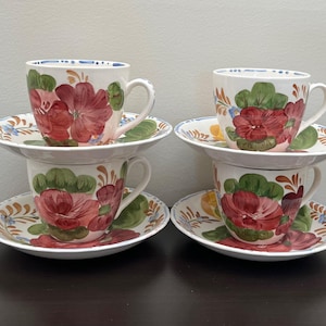 May include: Four white ceramic teacups and saucers. Each cup and saucer features a hand-painted floral design with red, green, and yellow accents. The cups have a rounded shape with a handle, and the saucers are round.