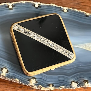 May include: A black and gold compact with an ornate design. The compact is made of metal and has a hinged lid.