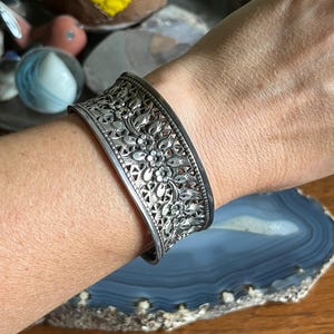 Puede incluir: Un brazalete de plata con intrincados diseños calados de flores y hojas. La ancha banda presenta un detallado calado, mostrando un patrón de flores y follaje. El brazalete se lleva en la muñeca, destacando la artesanía ornamentada.