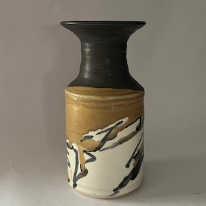 May include: A ceramic vase with a black rim and neck, transitioning to a tan band, and a white base. The vase features abstract black and tan brushstrokes. The vase has a wide mouth and a cylindrical body.