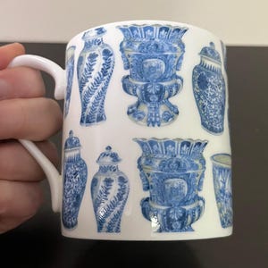 Puede incluir: Taza de cer&aacute;mica blanca con un patr&oacute;n repetido de jarrones y urnas decorativas azules y blancas. La taza tiene un asa curva y un acabado brillante. El dise&ntilde;o recuerda a la porcelana cl&aacute;sica.