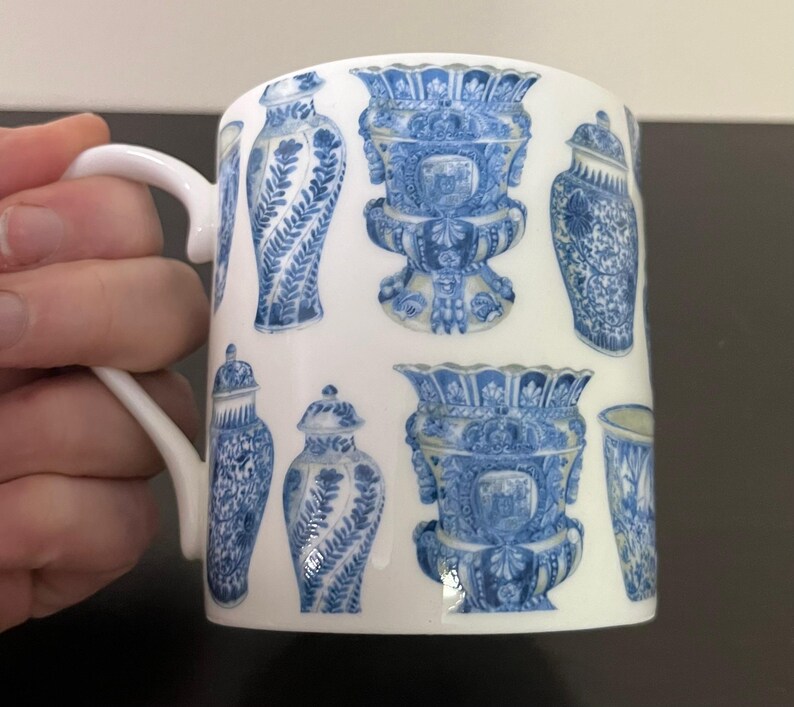 Puede incluir: Taza de cer&aacute;mica blanca con un patr&oacute;n repetido de jarrones y urnas decorativas azules y blancas. La taza tiene un asa curva y un acabado brillante. El dise&ntilde;o recuerda a la porcelana cl&aacute;sica.