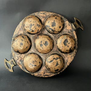May include: A circular metal pan with seven rounded cavities, finished in bronze with dark detailing. The pan features two decorative handles and a textured surface, giving it an antique or handcrafted look. The pan's diameter is approximately 20 cm.
