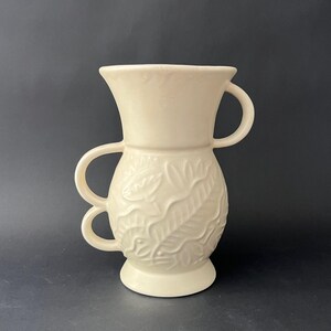May include: Cream-colored ceramic vase with two handles on each side. The vase features a detailed embossed leaf pattern around the middle and a flared top. The vase sits on a small, round base, and is set against a dark background.