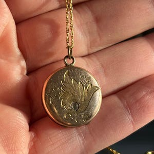 Può includere: Una collana con un ciondolo a forma di medaglione in oro con un designo floreale. Il medaglione è rotondo e ha una piccola pietra trasparente al centro.