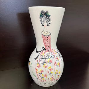 Può includere: Un vaso in ceramica bianca con un'illustrazione disegnata a mano di una donna in abito rosa. Il vaso ha una parte superiore svasata e una base bulbosa, con dettagli floreali e botanici. I capelli della donna sono acconciati in uno chignon con accenti verdi.