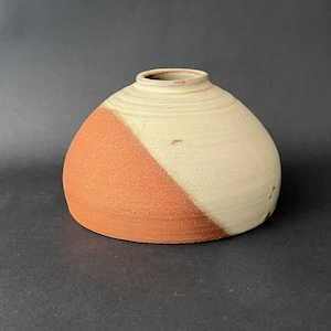 May include: A handmade ceramic vase with a rounded form and a small opening. The vase is divided diagonally, with one half in a warm terracotta hue and the other in a creamy off-white. The vase has a textured surface and a rustic appearance.