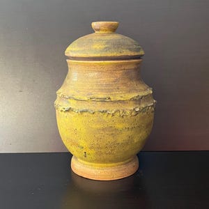Puede incluir: Un tarro de cerámica amarillo con tapa. El tarro tiene una superficie texturizada y una banda decorativa alrededor del centro. La tapa tiene un pequeño pomo en la parte superior. El tarro se asienta sobre una base redonda.