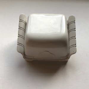 K&ouml;nnte beinhalten: Wei&szlig;e Keramik-Schmuckschatulle mit quadratischer Form und dekorativen Schn&ouml;rkeln an den Seiten. Die Schachtel hat ein erhabenes Design auf der Oberseite.