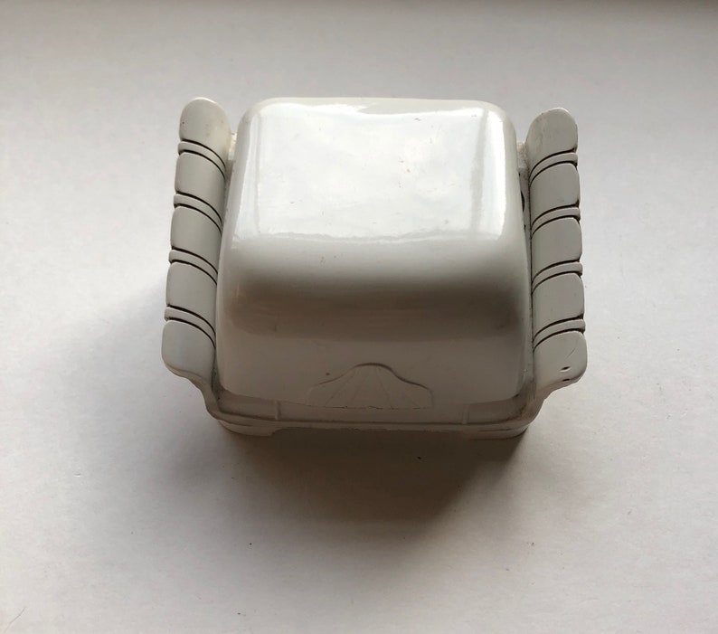 K&ouml;nnte beinhalten: Wei&szlig;e Keramik-Schmuckschatulle mit quadratischer Form und dekorativen Schn&ouml;rkeln an den Seiten. Die Schachtel hat ein erhabenes Design auf der Oberseite.