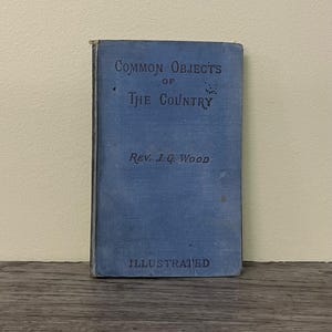 以下が含まれることがあります： Rev. J.G. Woodによる「Common Objects of the Country」というタイトルのヴィンテージ本。本は白い文字が入った青い表紙で、下部に「Illustrated」という文字があります。本は木製の表面に立っています。
