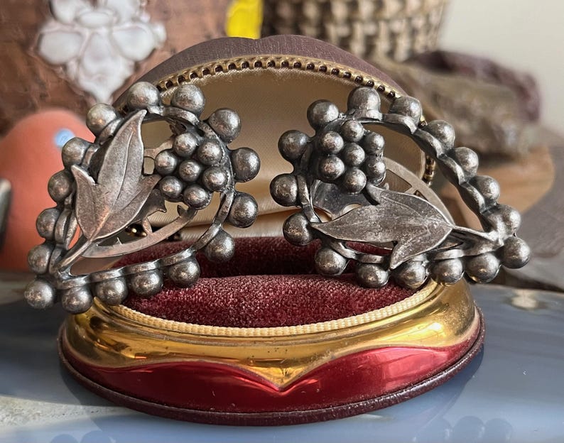 May include: A pair of silver tone earrings with a grapevine design. The earrings are in a velvet lined box.