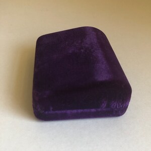 May include: A small, purple velvet box with a slightly curved top. The box is empty and appears to be made of a soft, plush material.
