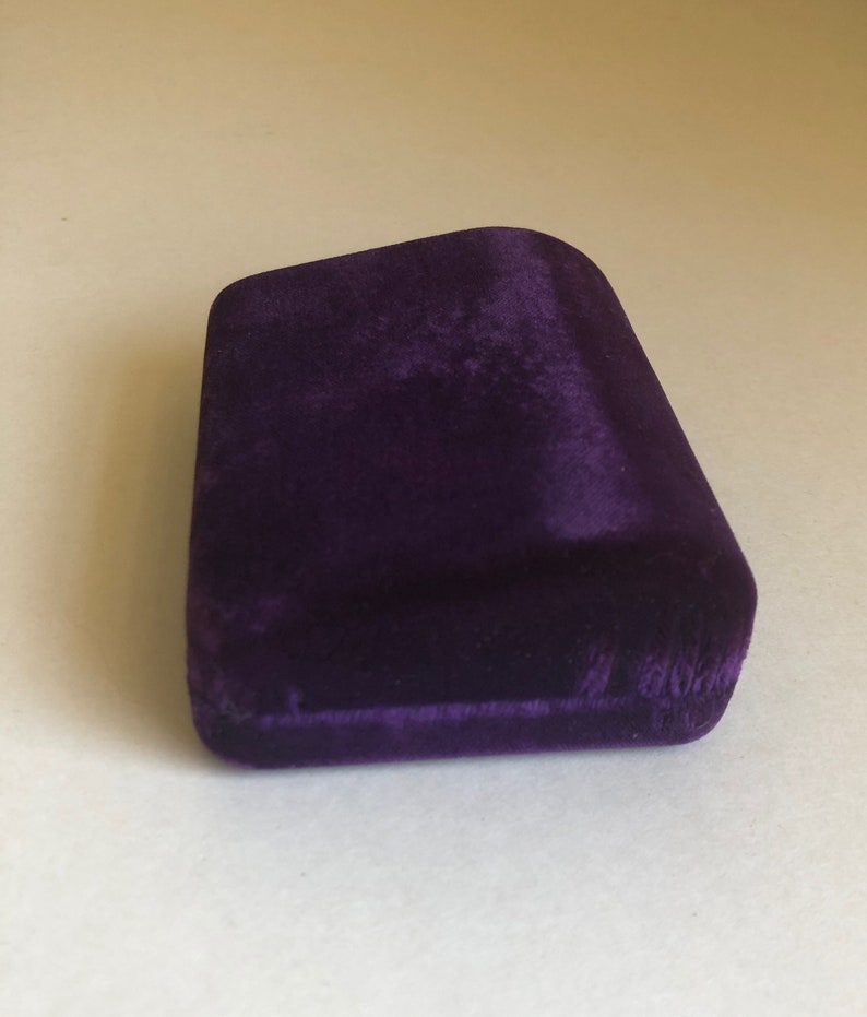 May include: A small, purple velvet box with a slightly curved top. The box is empty and appears to be made of a soft, plush material.