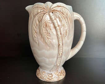 May include: A beige ceramic pitcher with a detailed palm tree design. The pitcher has a curved handle and a decorative base. The palm tree is in relief, with brown accents. The pitcher is likely for serving beverages.