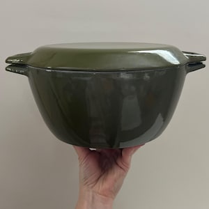 May include: An olive green enameled cast iron Dutch oven with a matching lid. The pot has two side handles. The cookware is designed for cooking and baking. The pot is held by a hand.