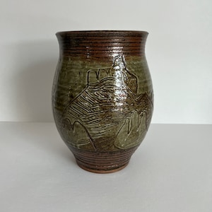 May include: A brown and green ceramic vase with a textured design of mountains and trees. The vase has a wide mouth and a rounded body.