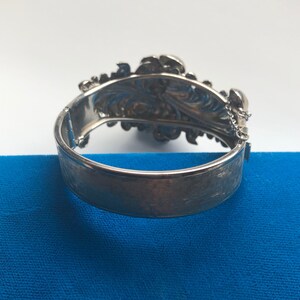K&ouml;nnte beinhalten: Ein silbernes Armband mit floralem Design und einem Kettenverschluss. Das Armband ist ge&ouml;ffnet und die R&uuml;ckseite ist sichtbar.