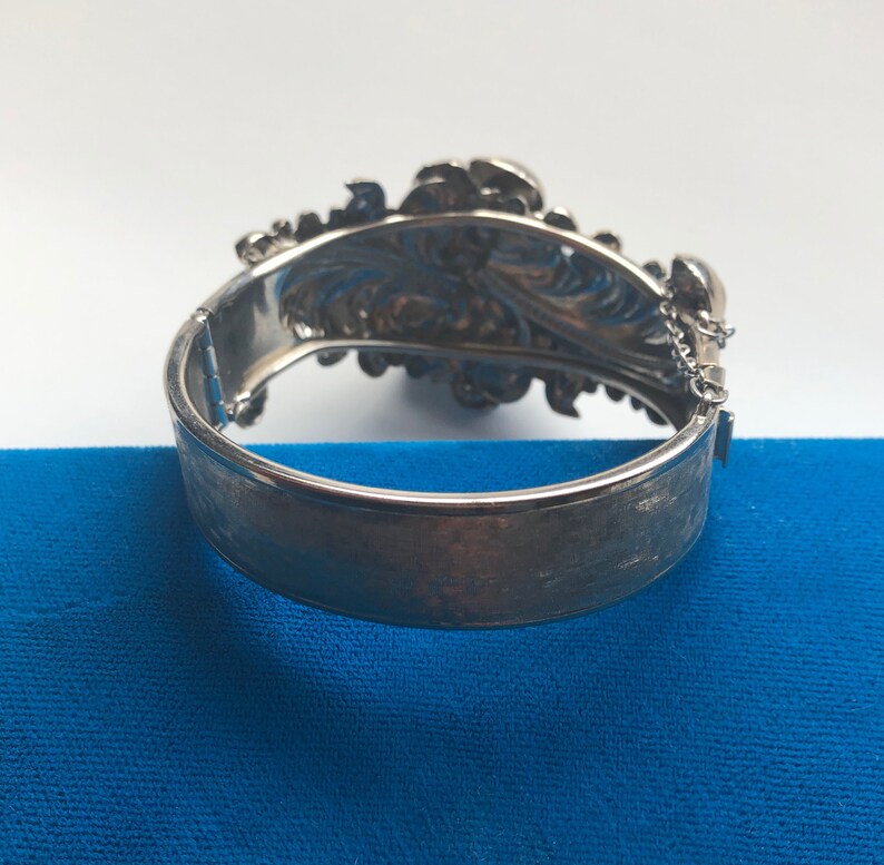 K&ouml;nnte beinhalten: Ein silbernes Armband mit floralem Design und einem Kettenverschluss. Das Armband ist ge&ouml;ffnet und die R&uuml;ckseite ist sichtbar.