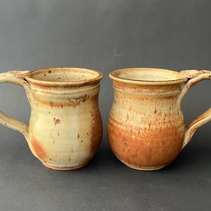 May include: Two handmade ceramic mugs with a rustic, speckled appearance. The mugs feature a cream-colored base that transitions to a warm orange-brown hue. Each mug has a curved handle and a decorative element where the handle meets the cup.