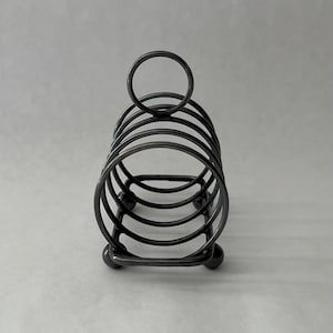 May include: A dark gray metal toast rack with a circular handle and a series of curved bars to hold toast. The rack has a rectangular base with small ball feet. The design is simple and functional.