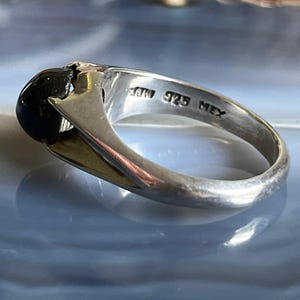 May include: Silver ring with a large, round, black stone. The ring has a polished finish and features the markings "925 MEX". The ring's band has a gold-colored accent.