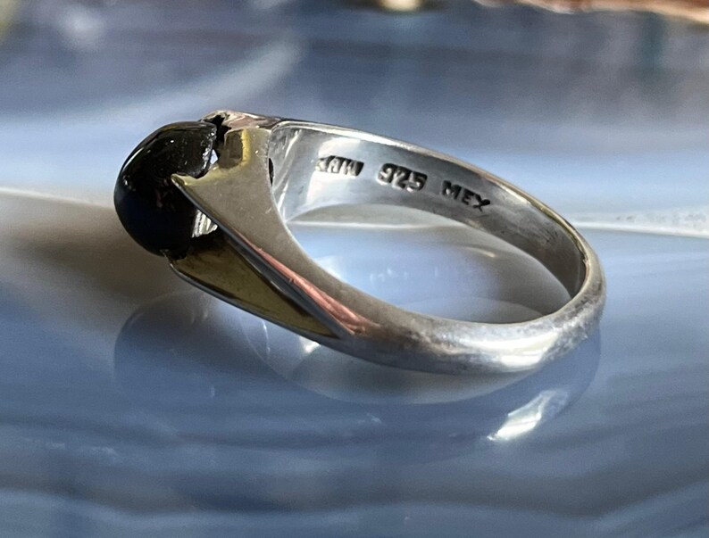 May include: Silver ring with a large, round, black stone. The ring has a polished finish and features the markings "925 MEX". The ring's band has a gold-colored accent.
