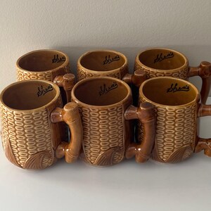 May include: Six brown ceramic mugs with a woven basket design and bamboo-style handles. Each mug has the word "Shea's" in a cursive font inside the rim. The mugs are arranged in a row on a white surface.