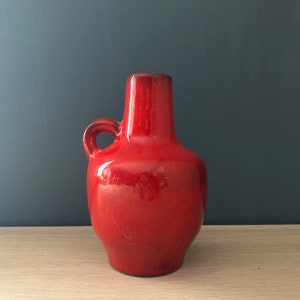 Puede incluir: Un jarrón de cerámica rojo con asa. El jarrón tiene un cuerpo redondeado y un cuello estrecho.