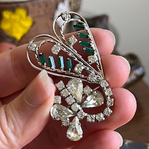 Puede incluir: Un broche de tono plateado con un diseño en forma de corazón con diamantes de imitación transparentes y verdes. El broche tiene un diseño delicado e intrincado.