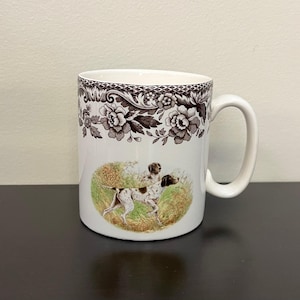 May include: White ceramic mug with a brown floral border and a detailed illustration of two dogs in a field. The dogs are brown and white, and the mug has a curved handle.