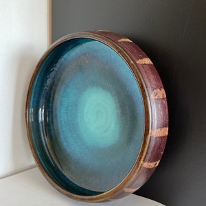 May include: A round ceramic bowl with a deep teal interior and a dark brown rim. The rim has a textured, brick-like pattern with lighter brown accents. The bowl is set against a white and black background.