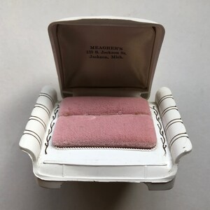 K&ouml;nnte beinhalten: Wei&szlig;e Ringbox mit einem rosafarbenen Samt-Innenraum. Die Box ist in Form einer kleinen Couch mit einer erh&ouml;hten R&uuml;ckenlehne und Armlehnen gestaltet. Der Deckel der Box ist ge&ouml;ffnet und die Innenseite des Deckels tr&auml;gt den Text "MEAGHER'S 133 S. Jackson St. Jackson, Mich."