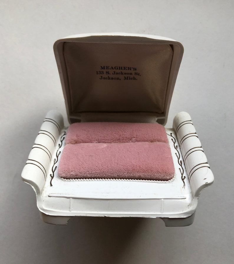 K&ouml;nnte beinhalten: Wei&szlig;e Ringbox mit einem rosafarbenen Samt-Innenraum. Die Box ist in Form einer kleinen Couch mit einer erh&ouml;hten R&uuml;ckenlehne und Armlehnen gestaltet. Der Deckel der Box ist ge&ouml;ffnet und die Innenseite des Deckels tr&auml;gt den Text "MEAGHER'S 133 S. Jackson St. Jackson, Mich."