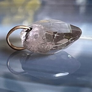 May include: A clear, faceted crystal pendant with a gold-colored bail. The crystal has a light brown hue and is partially translucent. The pendant is displayed on a reflective, blue-toned surface.