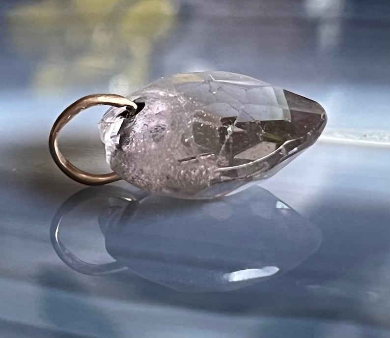 May include: A clear, faceted crystal pendant with a gold-colored bail. The crystal has a light brown hue and is partially translucent. The pendant is displayed on a reflective, blue-toned surface.