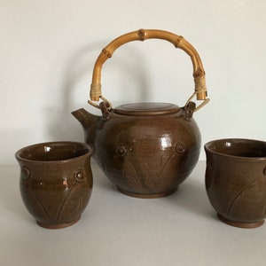 May include: A brown ceramic teapot with a bamboo handle and two matching brown ceramic cups. The teapot and cups have a floral design.