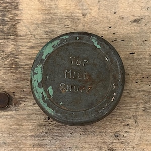 Op de afbeelding: Een ronde, antieke metalen container met de tekst "TOP MILL SNUFF" in reliëf op de bovenkant. De container heeft een verweerde, verouderde uitstraling met afbladderende groene verf langs de randen. De achtergrond is een verweerd houten oppervlak.