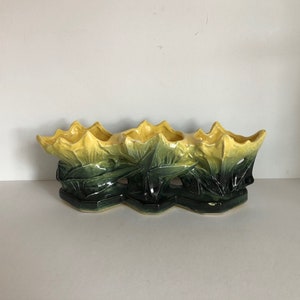 Puede incluir: Un macetero de cerámica amarillo y verde con tres aberturas en forma de flor. El macetero está decorado con un diseño floral.