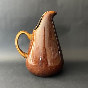 May include: A brown ceramic jug with a glossy finish. The jug has a curved handle and a spout for pouring. The teardrop-shaped body is a rich, warm brown colour. The jug is likely used for serving beverages.