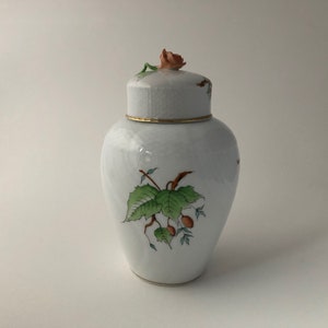 Peut inclure: Un pot en porcelaine blanche avec un motif floral. Le pot a un couvercle avec une petite rose rose sur le dessus. Le pot est décoré de feuilles vertes et de baies brunes.