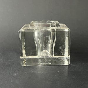 May include: A clear glass inkwell with a square base and a central cylindrical ink reservoir. The inkwell is made of thick, transparent glass. The design is simple and functional, suitable for a desk or writing area. The inkwell is made of thick, transparent glass.