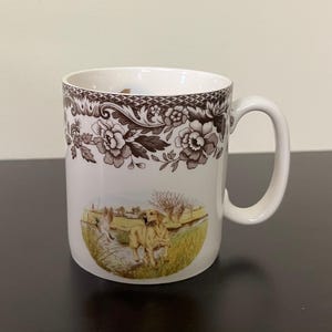 May include: White ceramic mug with a brown floral pattern and a rural scene featuring a golden retriever. The mug has a curved handle. The background is neutral.